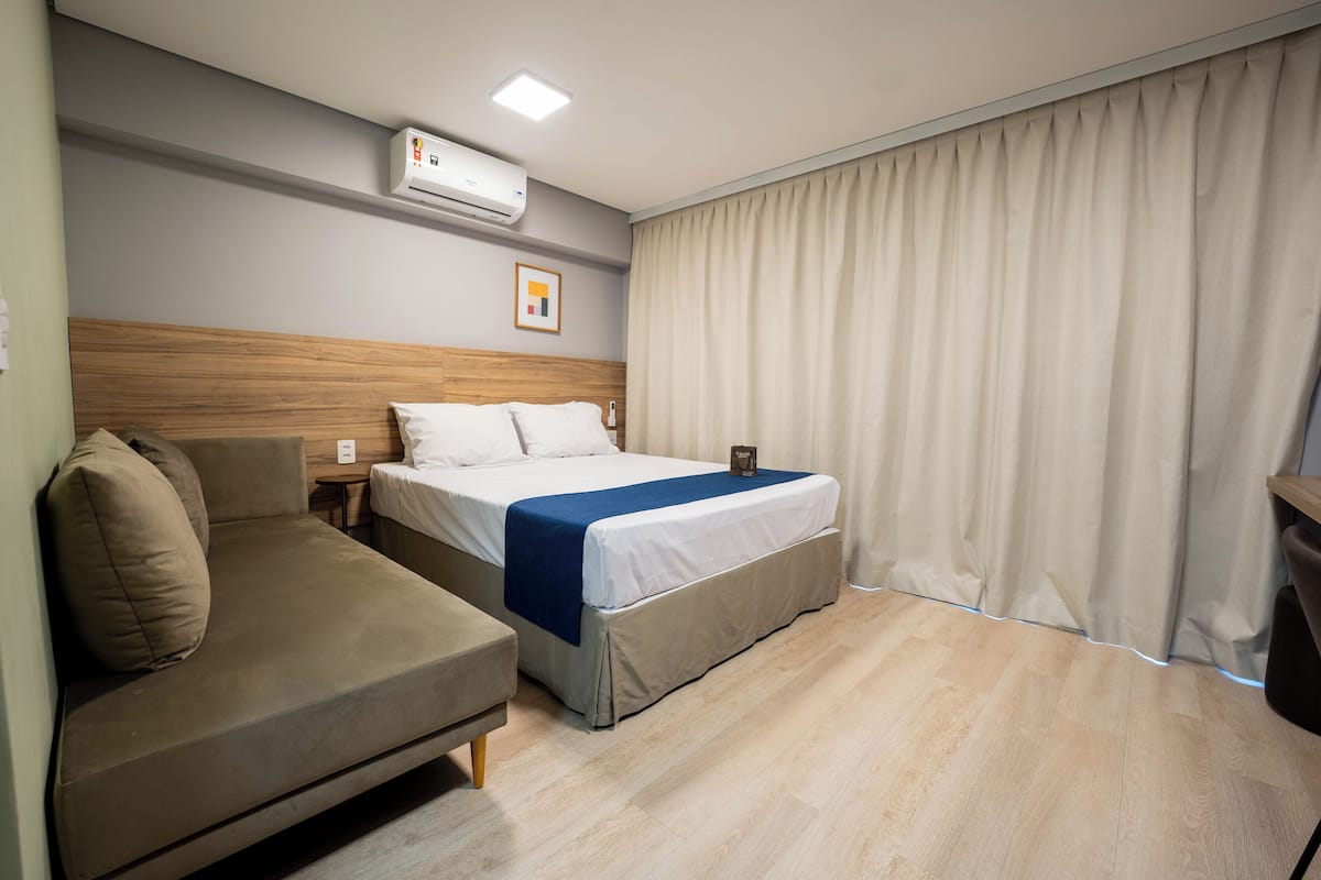 Una habitación de hotel con cama, sofá, unidad de aire acondicionado, mesita de noche y cortina.