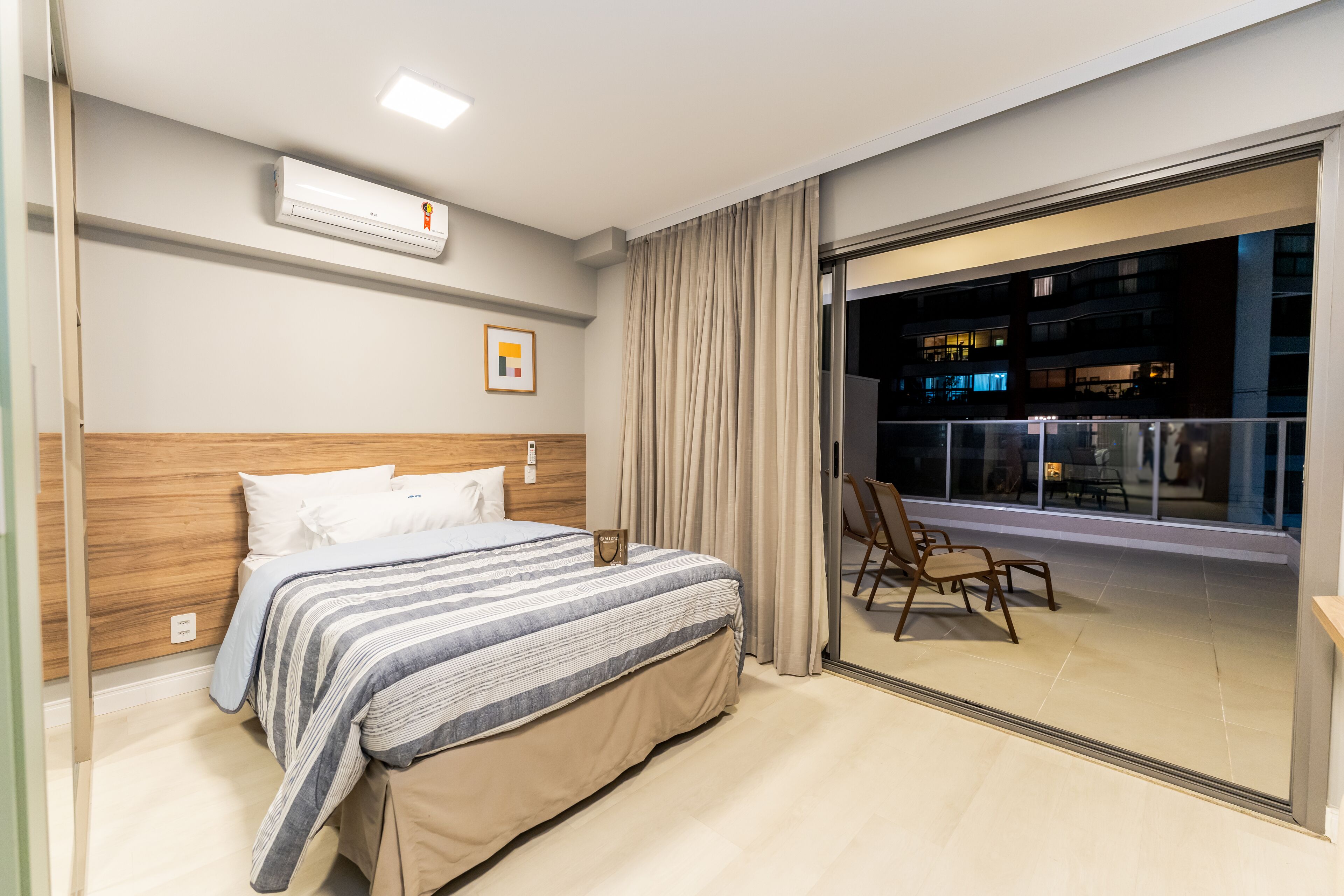 Una habitación de hotel moderna con una cama grande, cabecera de madera, puerta corrediza de vidrio a un balcón y vistas de la ciudad de noche.