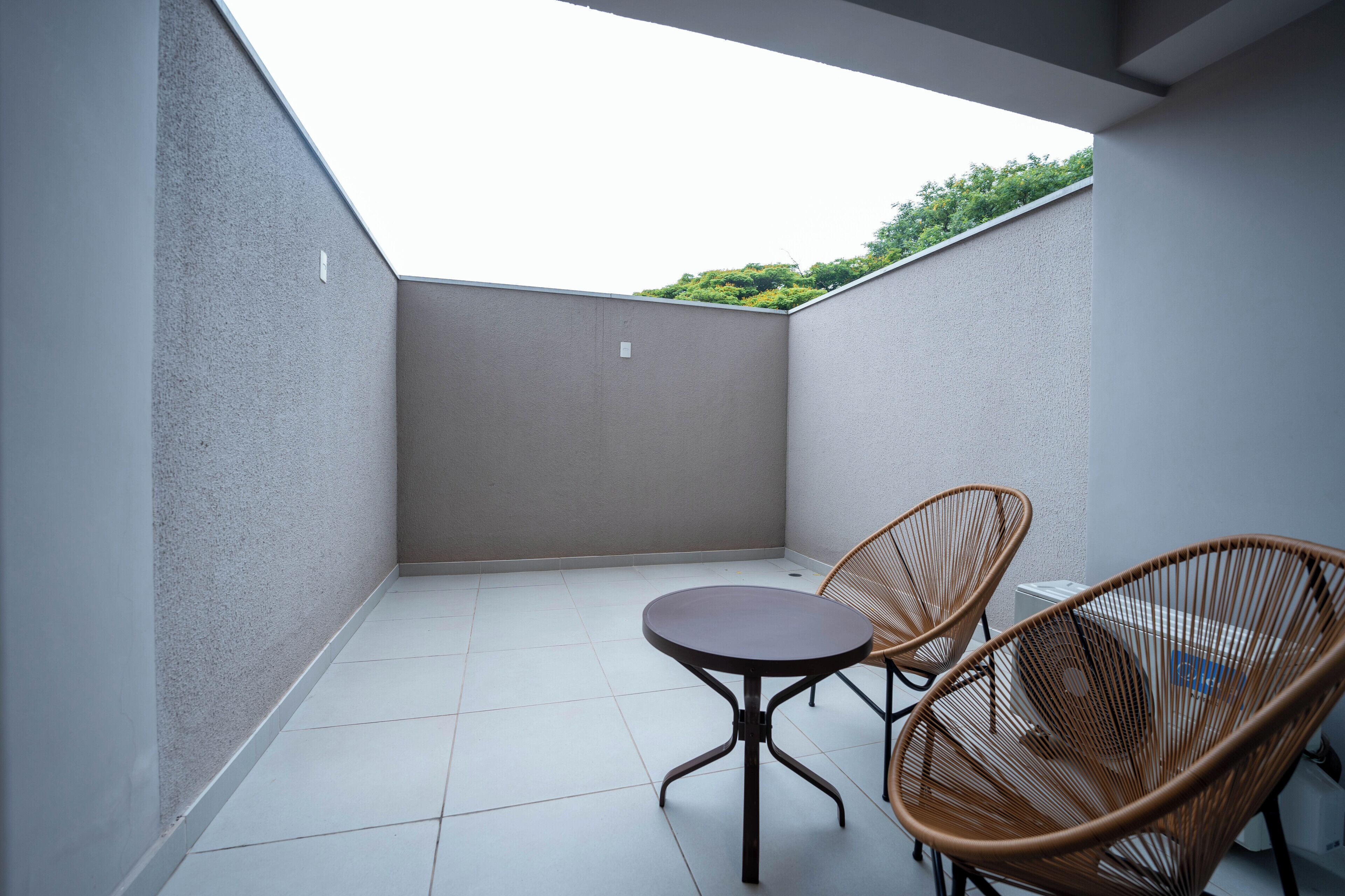 A small, enclosed balcony with two wicker chairs and a round table, featuring a tiled floor and a view of greenery in the background.