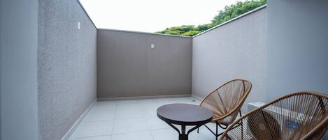 Un piccolo balcone privato con due sedie in vimini e un tavolo rotondo, pavimento piastrellato e vista su piante nel verde sullo sfondo.