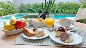 A breakfast setup with fruits, pastries, and beverages on a table by a poolside with a view of a building and greenery. - Apartamentos Studio Allure Moema (São Paulo)