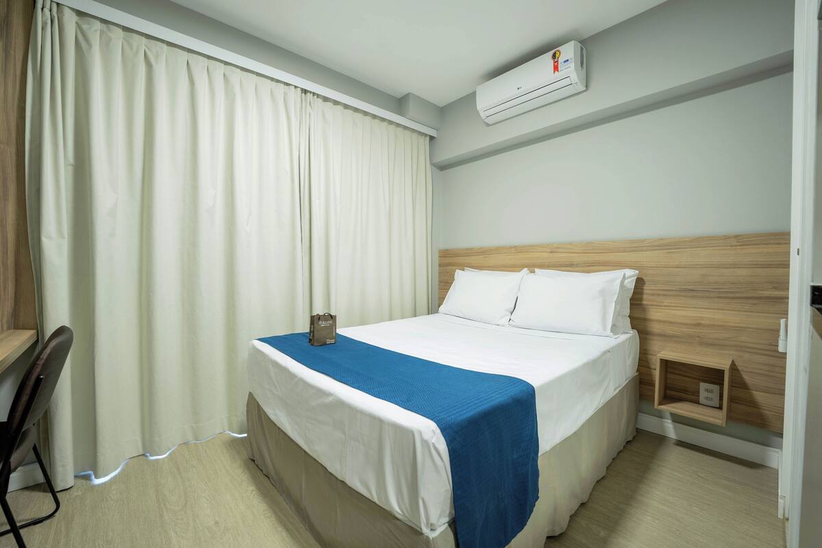 Habitación de hotel con cama, escritorio, silla, mesita de noche y aire acondicionado.