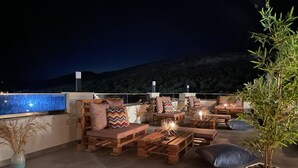 Eine Dachterrasse mit Holzmöbeln, einem Topfpflanzen und Blick auf die Berge bei Nacht.