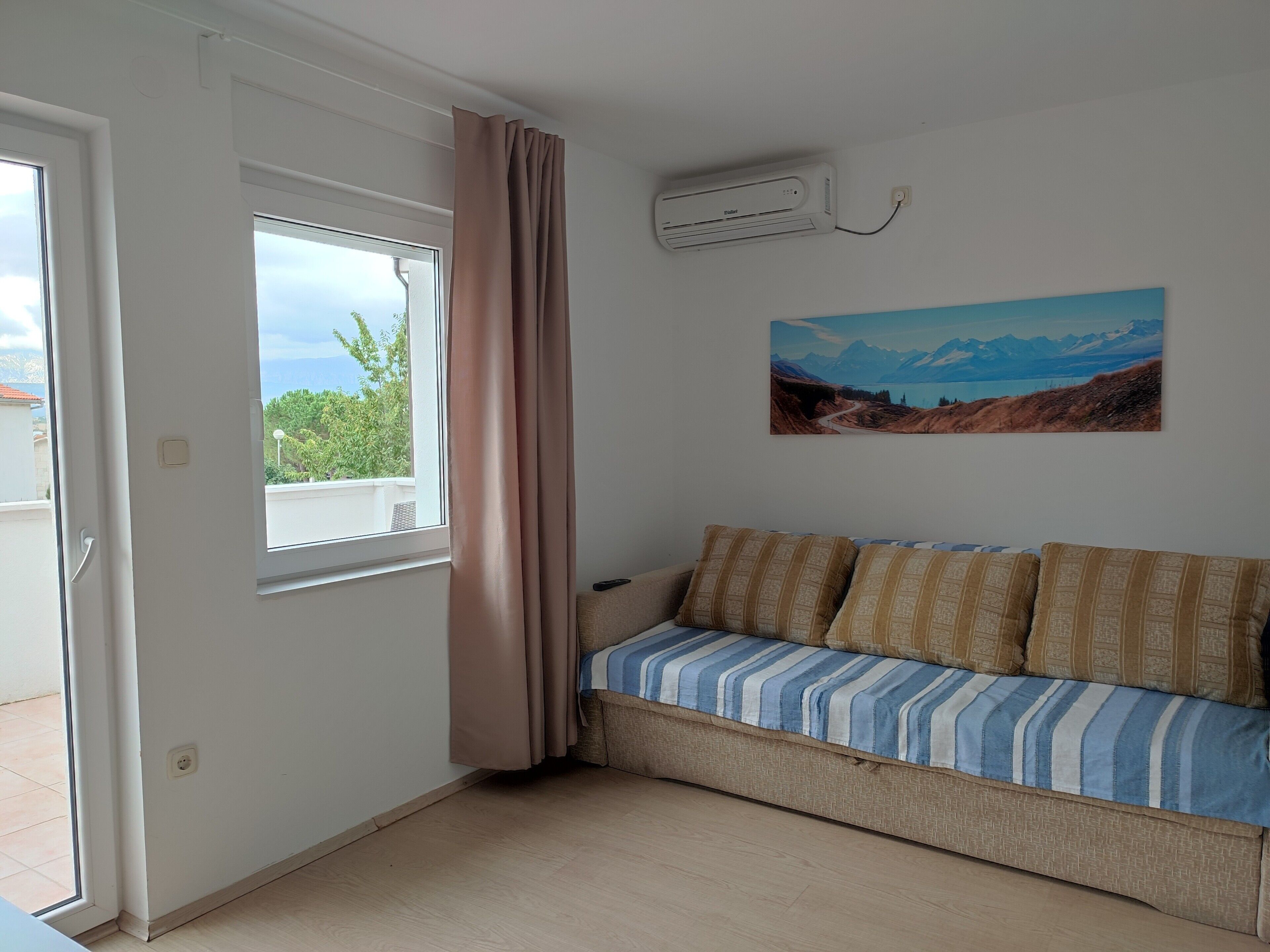Una habitació amb un sofà beix, una finestra amb vista a vegetació i un aparell de aire condicionat a la paret.
