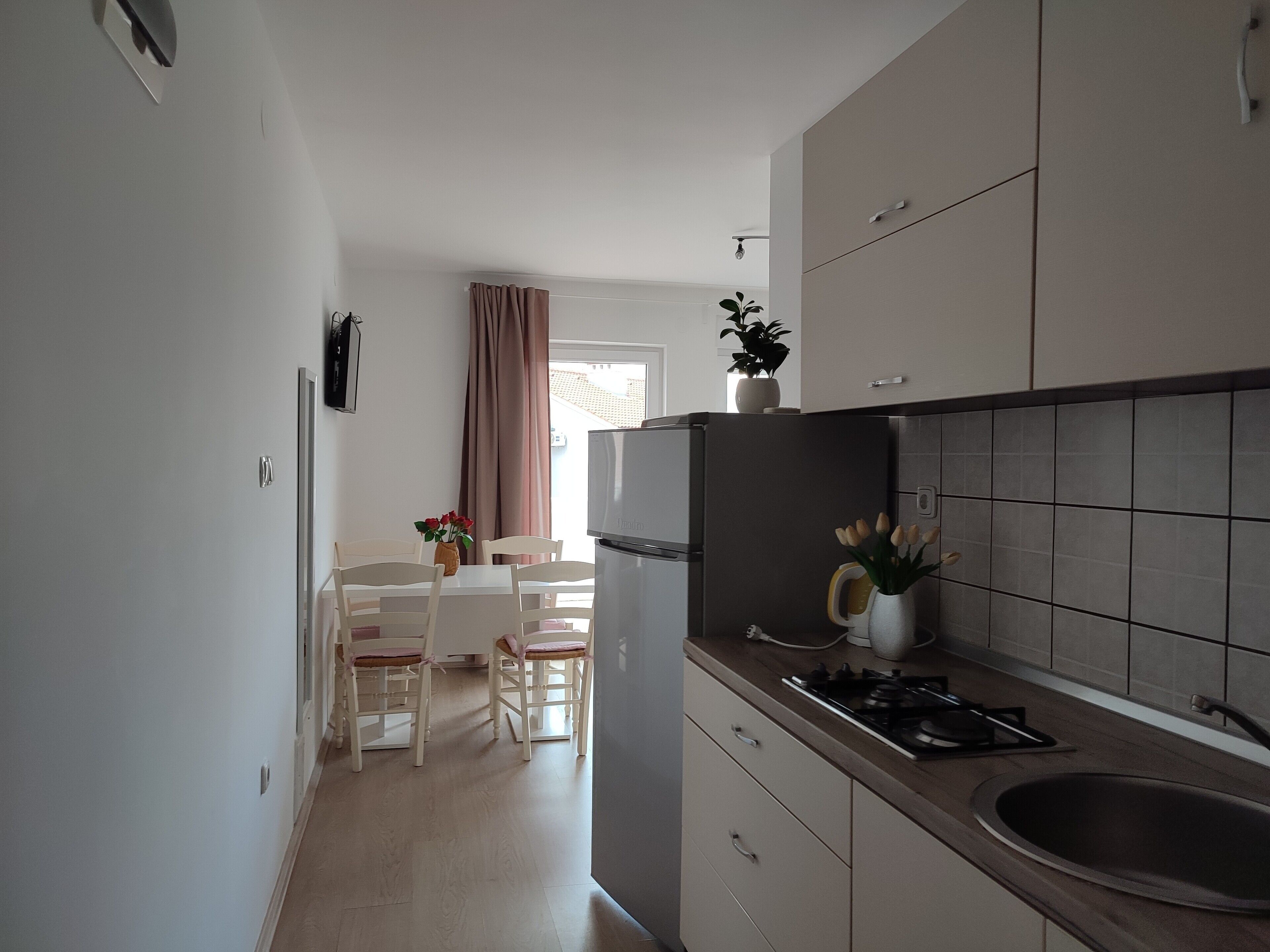 Een kleine keuken met een koelkast, fornuis en spoelbak. Er is een zithoek met een tafel en stoelen. Op de achtergrond is een raam met gordijnen te zien.