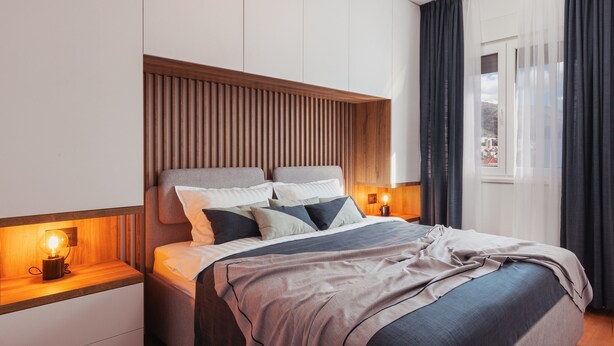 Une chambre d’hôtel moderne avec un grand lit, une tête de lit en bois, des lampes de chevet et une vue sur la ville à travers de fines rideaux.