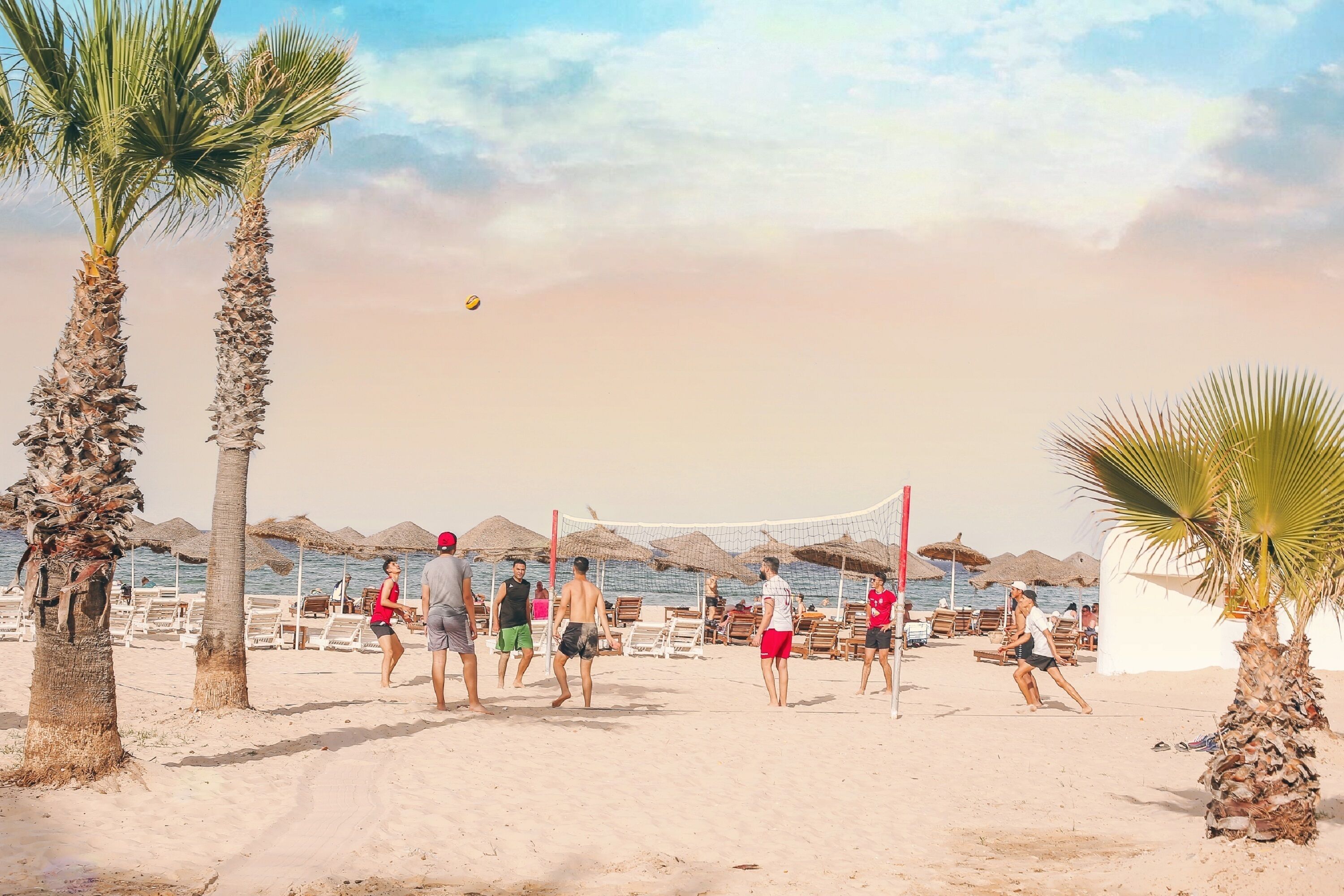 A beach volleyball game with players and spectators, palm trees, and beach umbrellas.