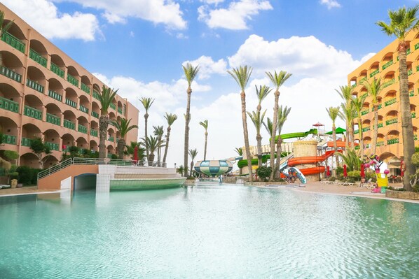 Zona de piscina de hotel con tobogán de agua, palmeras y un edificio de varios pisos con balcones.