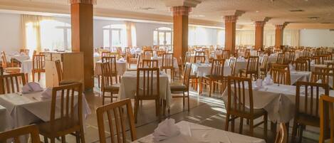 A large banquet hall with rows of tables and chairs, high ceilings with patterned columns, and large windows allowing natural light.