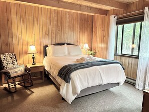Une chambre à coucher aux murs lambrissés, avec un lit, deux lampes de chevet, un fauteuil à bascule et une fenêtre donnant sur la forêt.