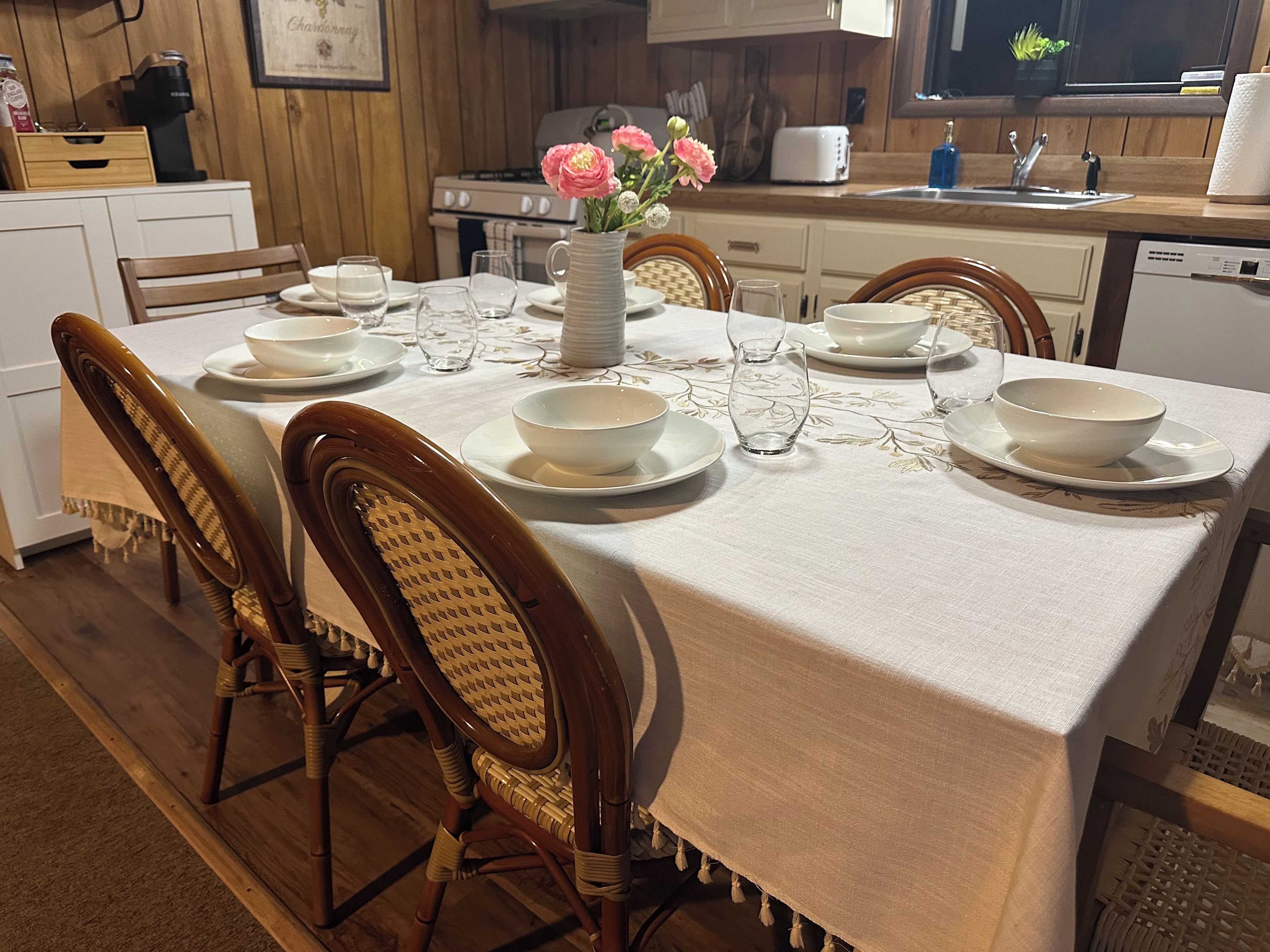 Une table à manger dressée avec de l’assiette, des bols et des verres blancs, entourée de chaises en osier.