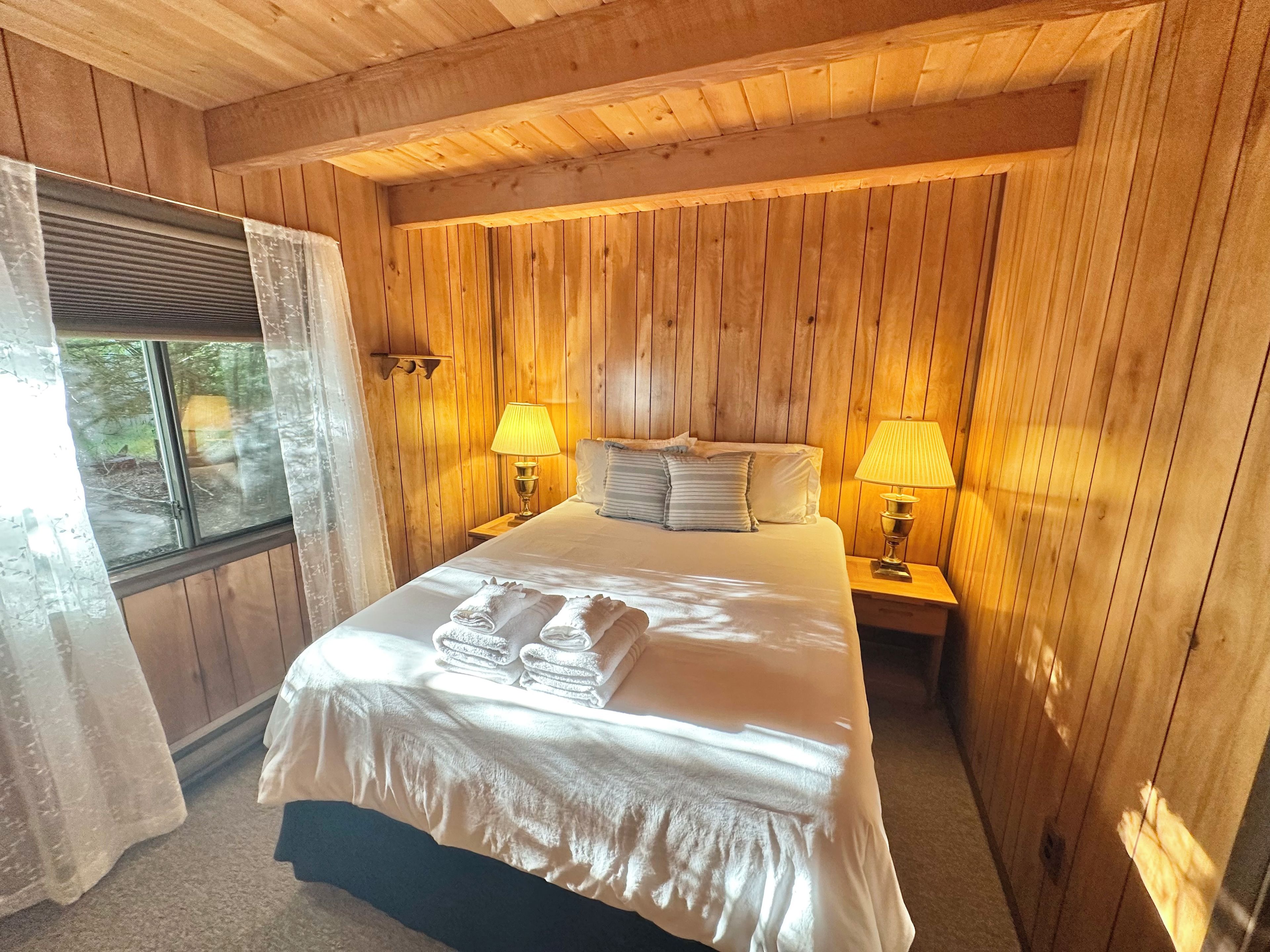 A wooden-paneled room with a bed, two bedside lamps, and a window with curtains.