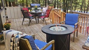 Une terrasse en bois avec une table ronde et des chaises, un foyer et un espace barbecue.