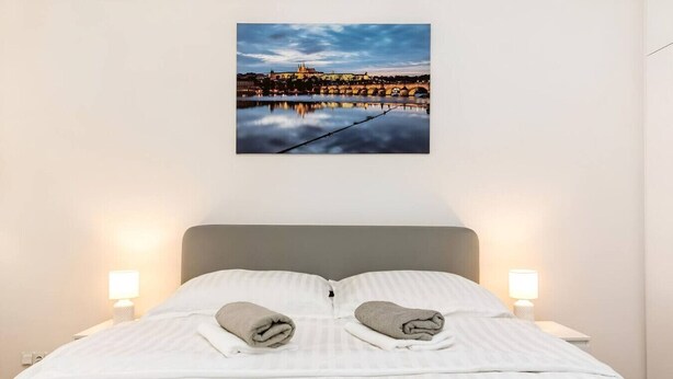 Una cama bien hecha con dos toallas, un cuadro enmarcado de un paisaje urbano y dos lámparas de noche.