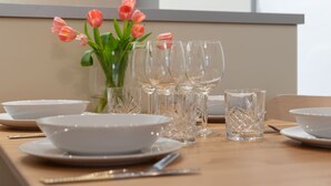 Una mesa de comedor preparada con platos, tazones, cubiertos y vasos, con un jarrón de tulipanes rosas al fondo.