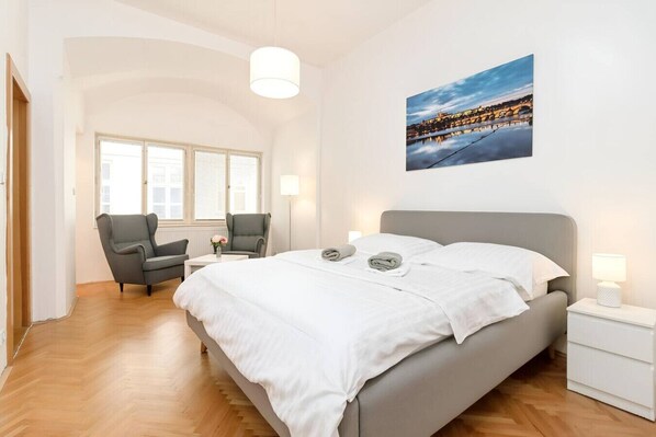 Une chambre d’hôtel avec un lit, deux fauteuils, une petite table, une table de chevet, une lampe, un tableau et une vue sur le paysage urbain.