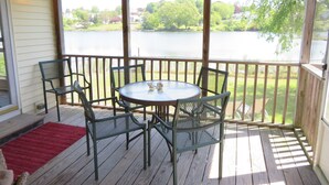 A covered outdoor deck with a round table and chairs, overlooking a lake with trees and houses in the background. - Waterfront Cottage within walking distance to the Beach. A dog-friendly property (Waterford)