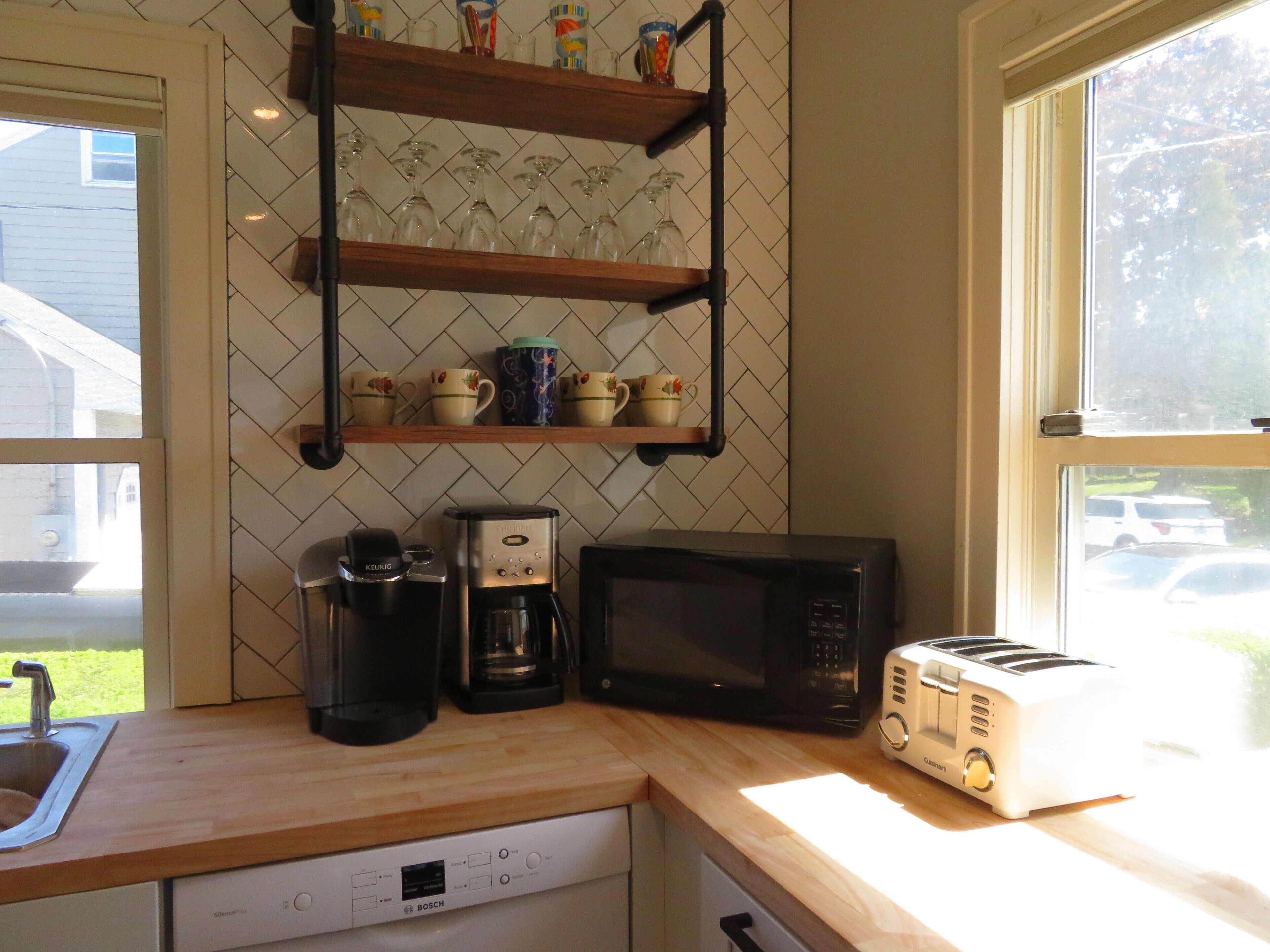 A kitchen with a Bosch dishwasher, a Keurig coffee maker, a microwave, and a toaster. There are shelves above holding cups and a window to the right.
