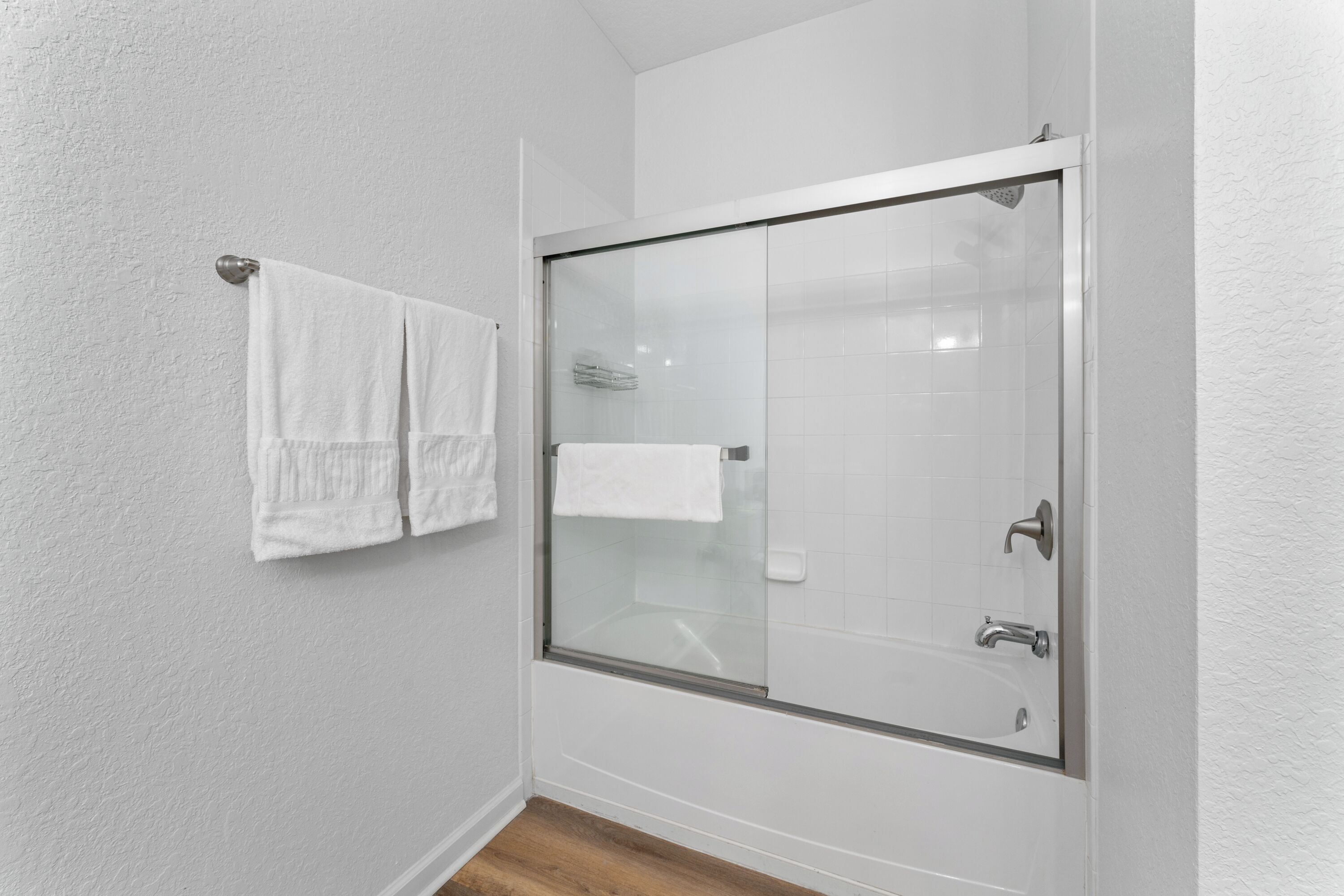 A bathroom with a glass-enclosed shower, white tiled walls, and a wooden floor.