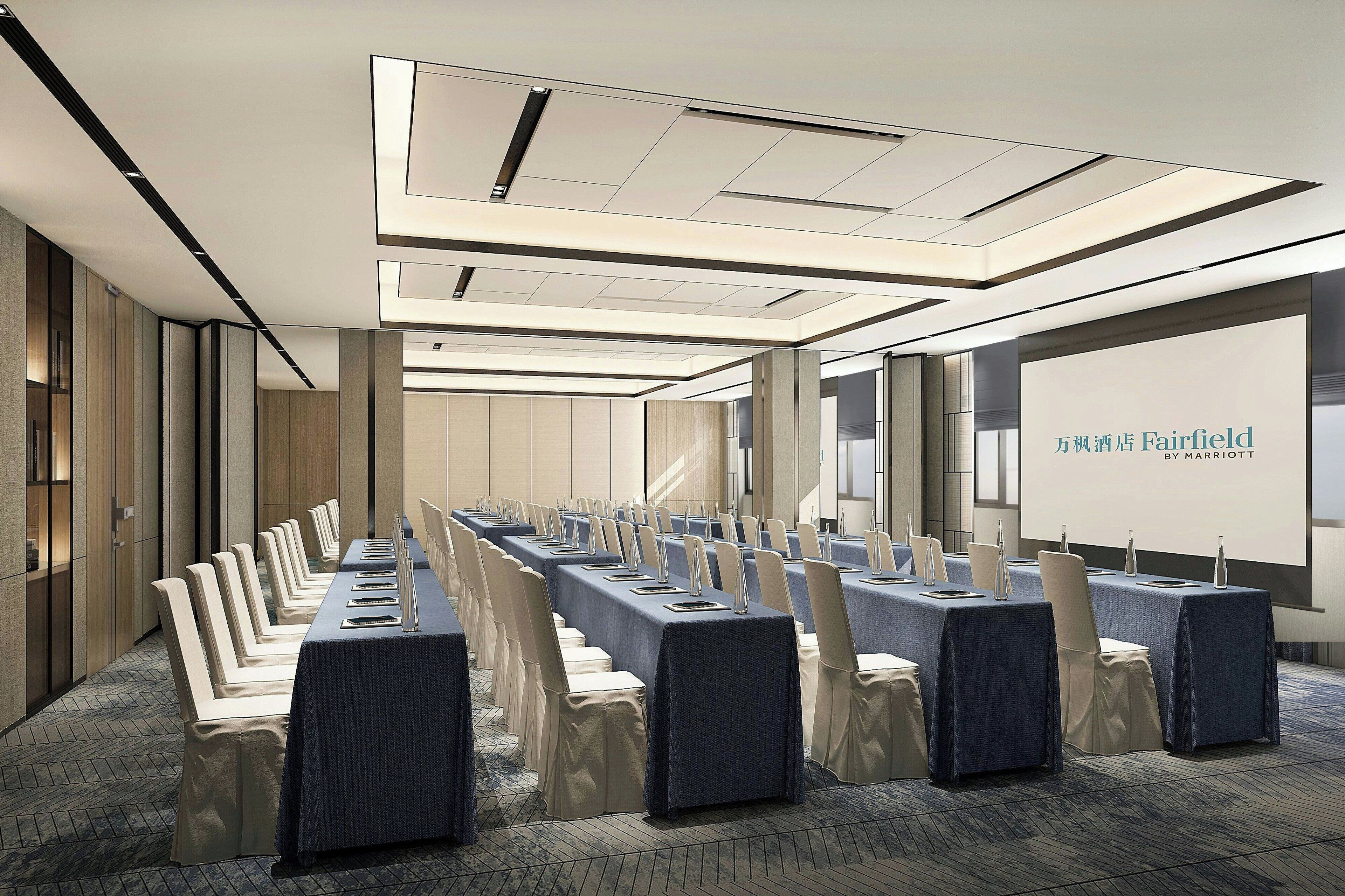 Un salón de conferencias con largas mesas, sillas y una pantalla de proyección que muestra 'Fairfield by Marriott'.