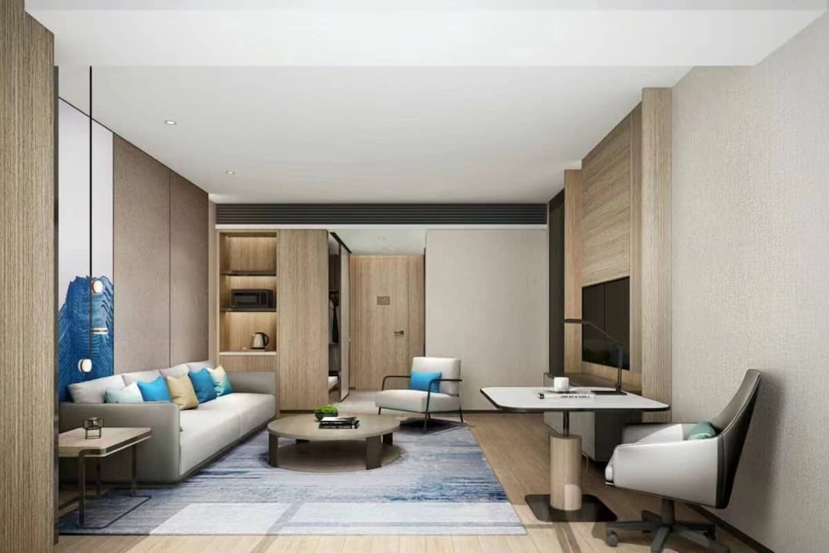 A modern hotel room with a sofa, coffee table, armchair, and a desk with a chair.