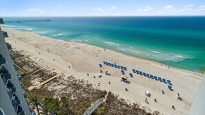 A beach with a pier in the background, a sandy shore, and a row of blue umbrellas. - Sterling Breeze 1205 - Gulf Front Condo (Panama City Beach)