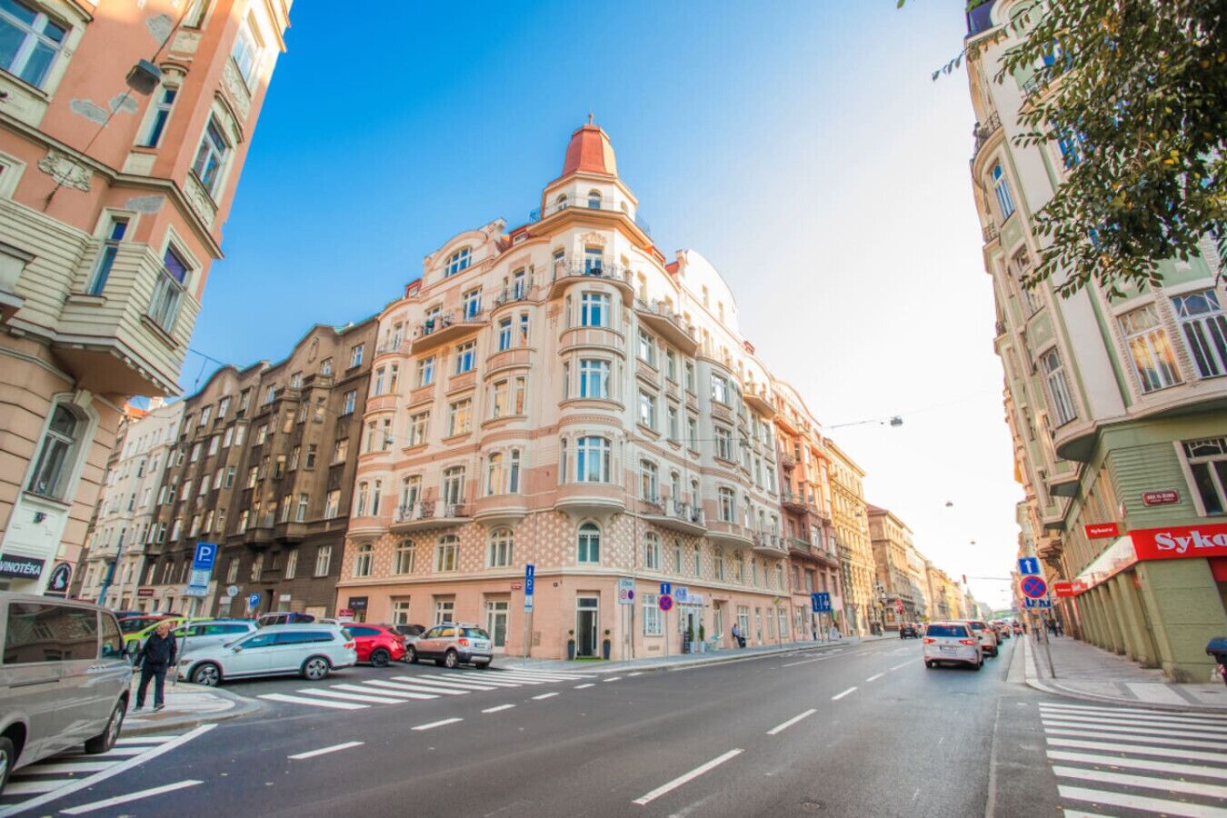 Ein mehrstöckiges Gebäude mit Hauseingang in der Ecke, einer Fußgängerampel und parkenden Autos an der Straße.