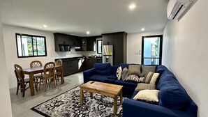 Ein modernes Wohnzimmer mit einem blauen Sofa, einem Holzespeise Tisch und Stühlen. Im Hintergrund ist eine Küche mit dunklen Schränken und einem Fenster zu sehen.