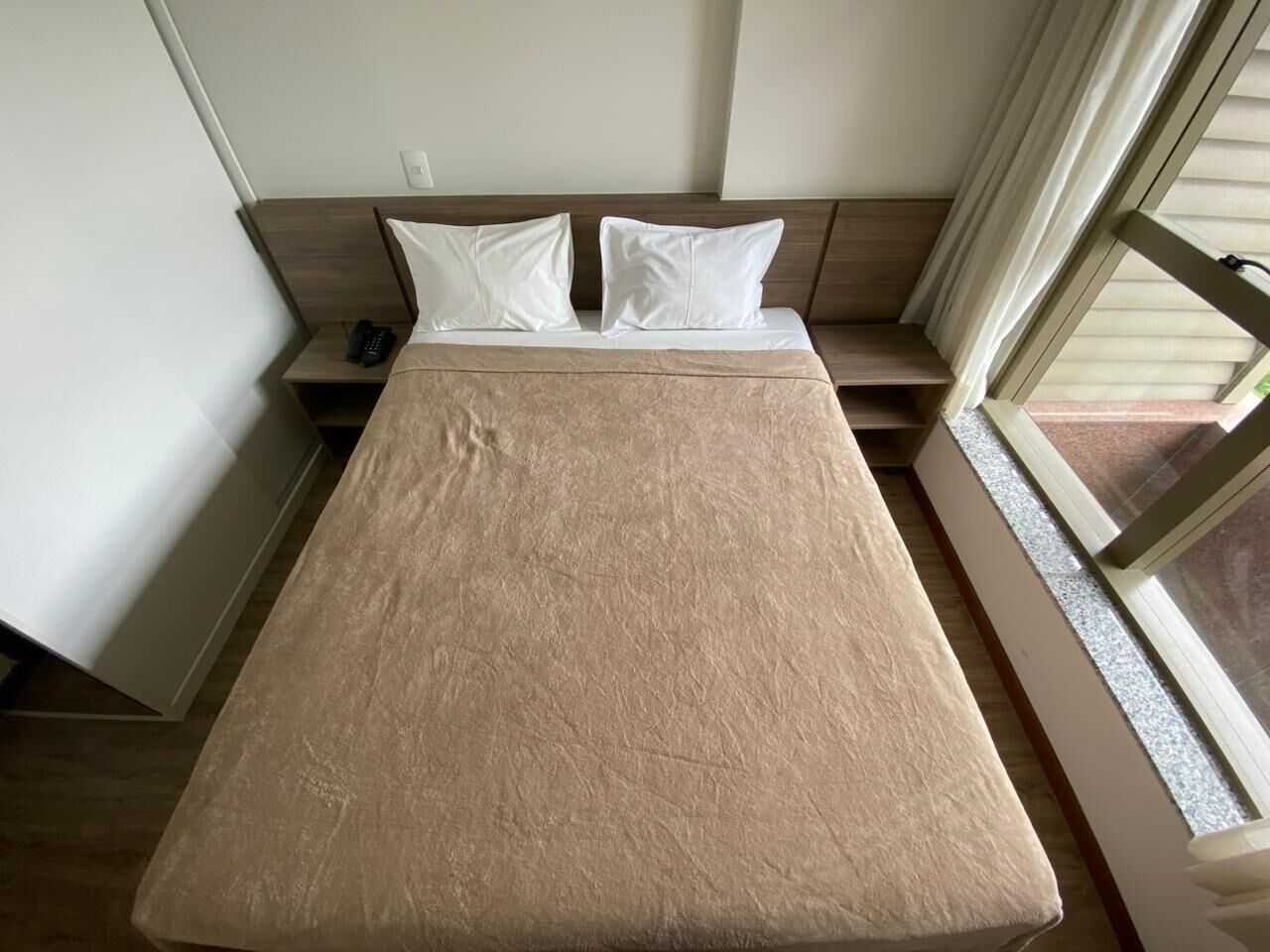Uma cama de solteiro com um espalha cama bege, duas almofadas brancas e uma mesa de cabeceira com um telefone.