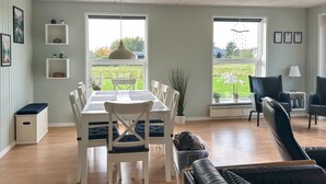 Ein Essbereich mit einem weißen Tisch und Stühlen, einer Topfpflanze und Blick auf eine grüne Landschaft durch große Fenster.