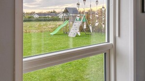 Blick durch ein Fenster auf eine grüne Außenanlage mit einem Spielplatz, darunter eine Rutsche und Schaukeln, und einem Gebäude im Hintergrund.