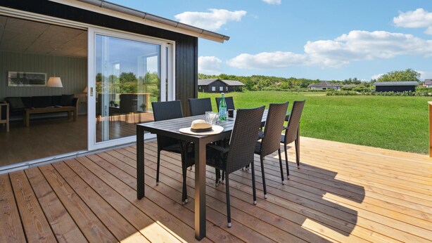 Eine Terrass mit Tisch und Stühlen, eine Glastür, die zu einem Raum führt, und ein Blick auf ein grünes Feld und Häuser in der Ferne.