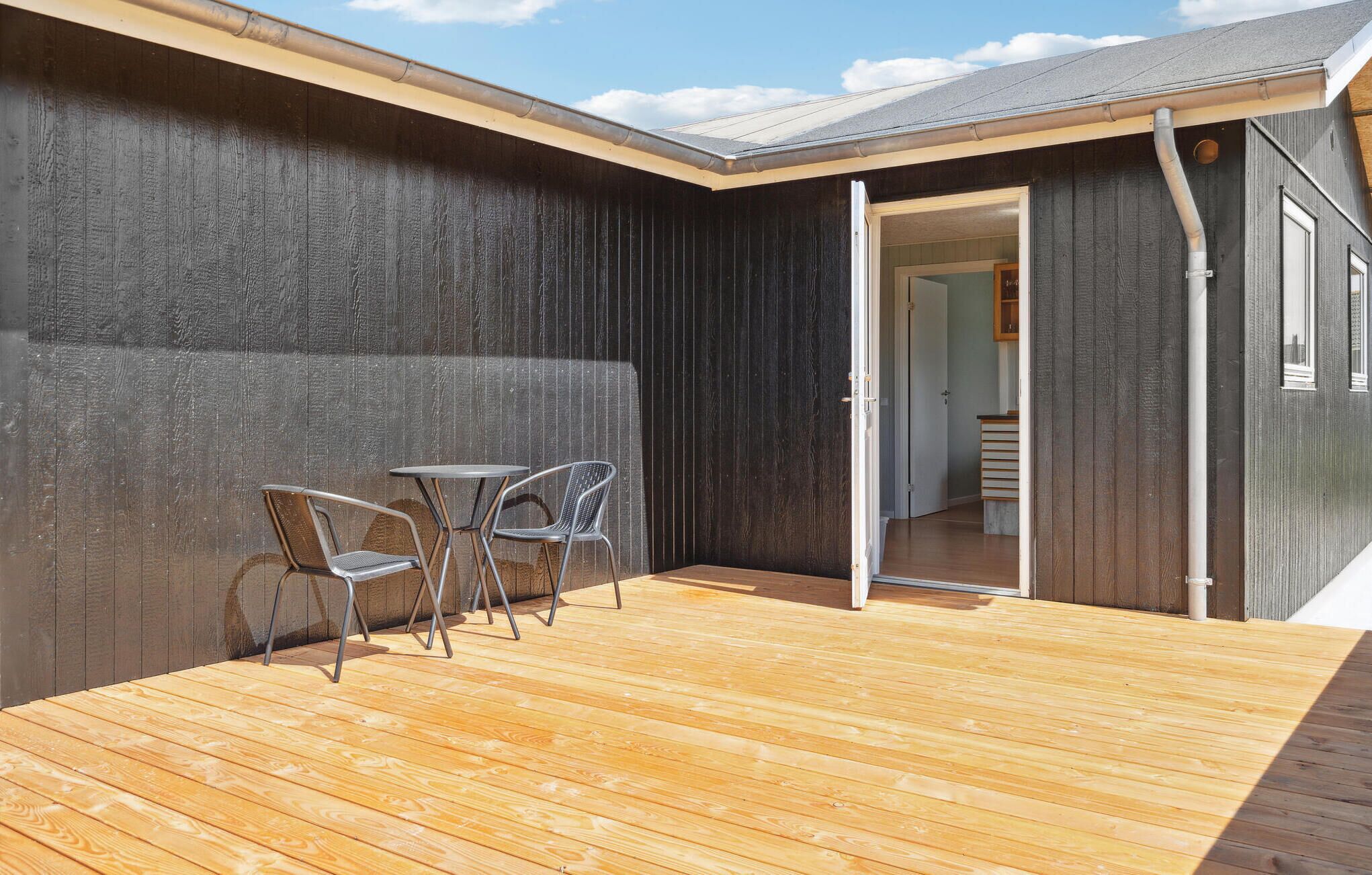 Une terrasse en bois avec deux chaises en métal et une table, un mur en bois foncé et une porte donnant sur un espace intérieur.
