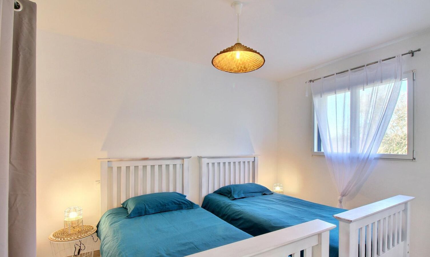 Een slaapkamer met twee bedden, een hanglamp, een nachtkastje met een lamp en een raam met dunne gordijnen.