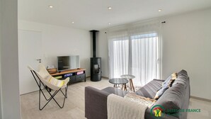 A modern living room with a sofa, a TV on a wooden stand, a small table, and a black wood-burning stove. - Le 26 - Erdeven - Gîte (ERDEVEN)