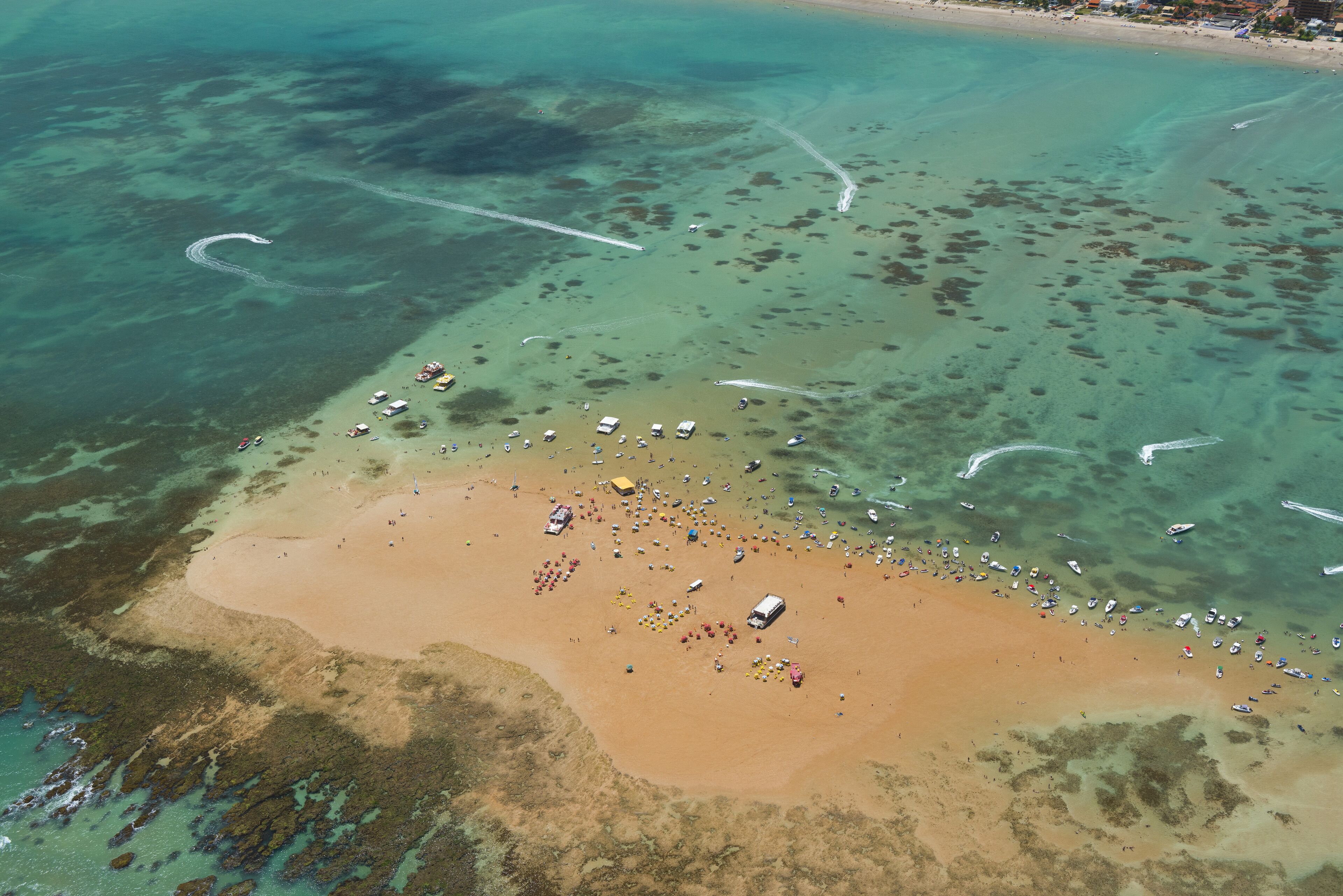 An aerial view of a beach with clear shallow waters, boats, and a sandy shore with umbrellas and people.