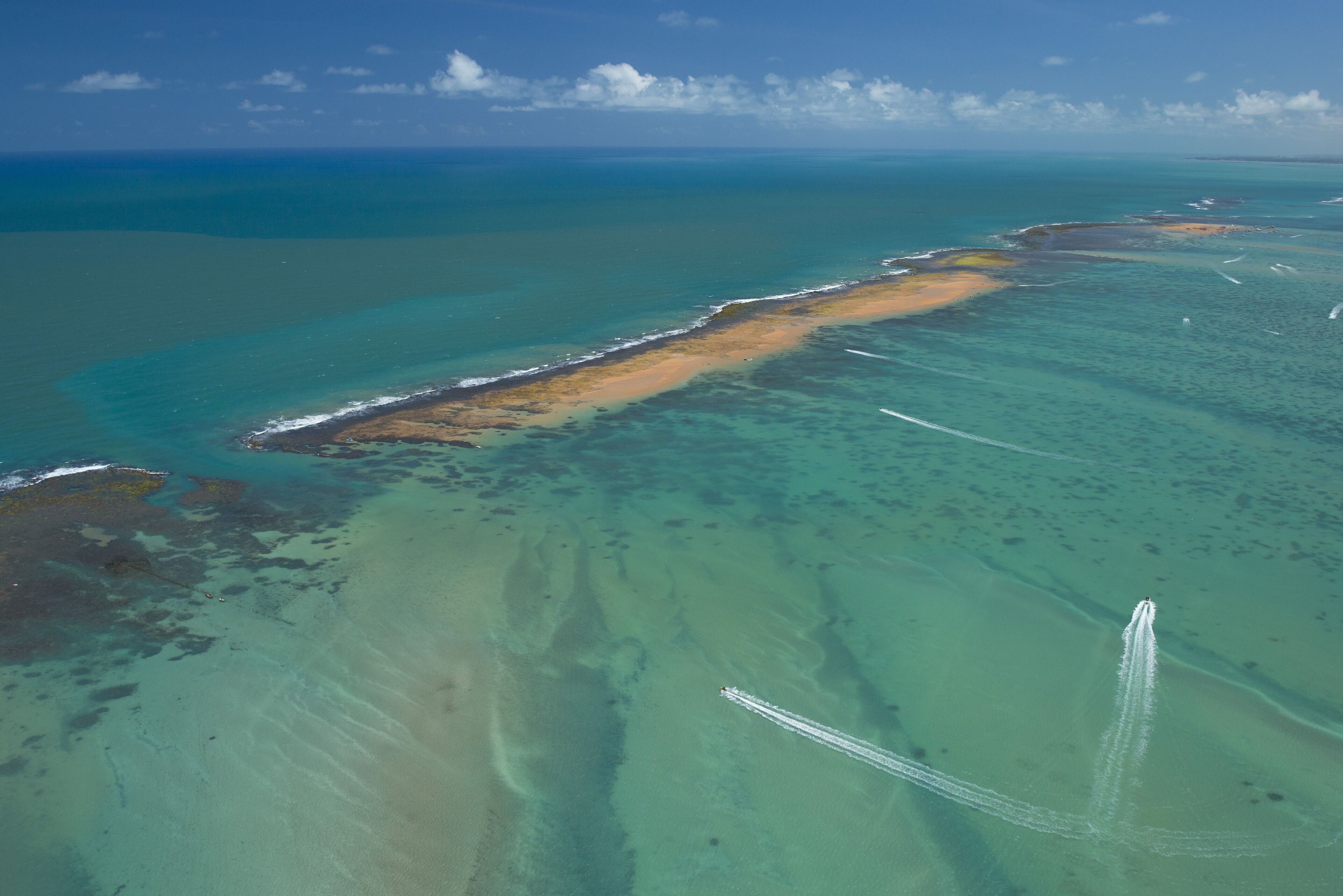 An aerial view of a coastal area with clear turquoise waters, sandy beaches, and a visible boat trail.
