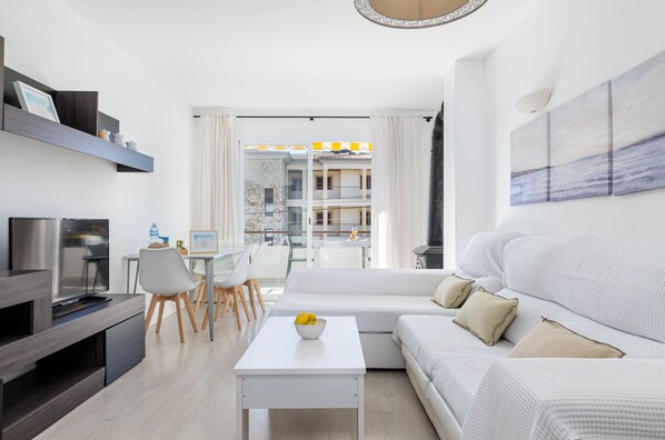 A modern living room with a sofa, a coffee table, and a TV. There is a dining area with chairs and a table. - YourHouse Llimonera, beach flat in Puerto de Pollença (Port de Pollença)