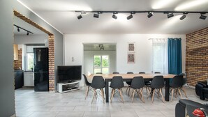 A modern dining area with a long table and chairs, a kitchen with a refrigerator and stove, and a TV on a stand.