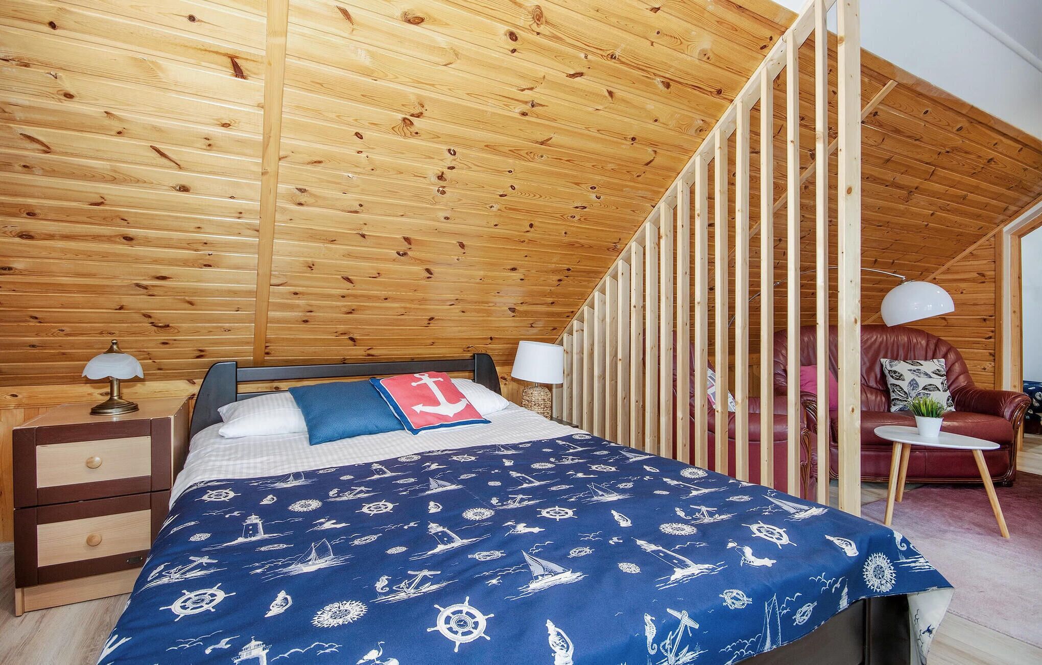 A bedroom with a wooden ceiling, a bed with a nautical-themed bedspread, a nightstand, a lamp, and a red leather sofa.