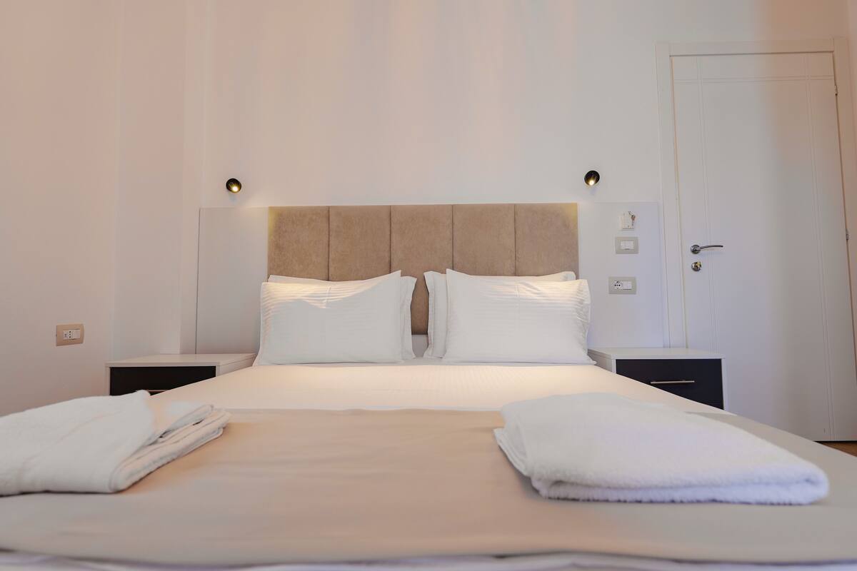 A neatly made bed with white linens and pillows, two bedside tables with lamps, and a door to the right.