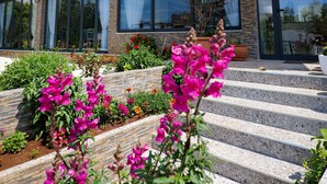 A modern two-story house with a balcony, large windows, and a garden with flowers.