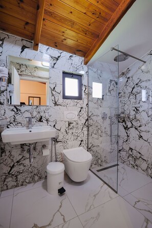 Un bany amb paret de marbre, un lavabo, un vàter i una cabina de dutxa de vidre.