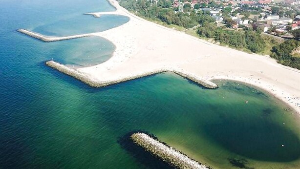 Vue aérienne d’une zone côtière avec une plage de sable, des eaux d’un bleu limpide et une petite île surmontée d’un promontoire rocheux.