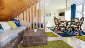 Un salon chaleureux avec un plafond en bois, un canapé agrémenté de coussins à motifs, une table basse en osier et un coin repas avec des chaises et une table en osier.