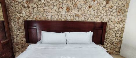 Habitación de hotel con pared de piedra, cabecero de madera, ropa de cama blanca y suelo estampado.