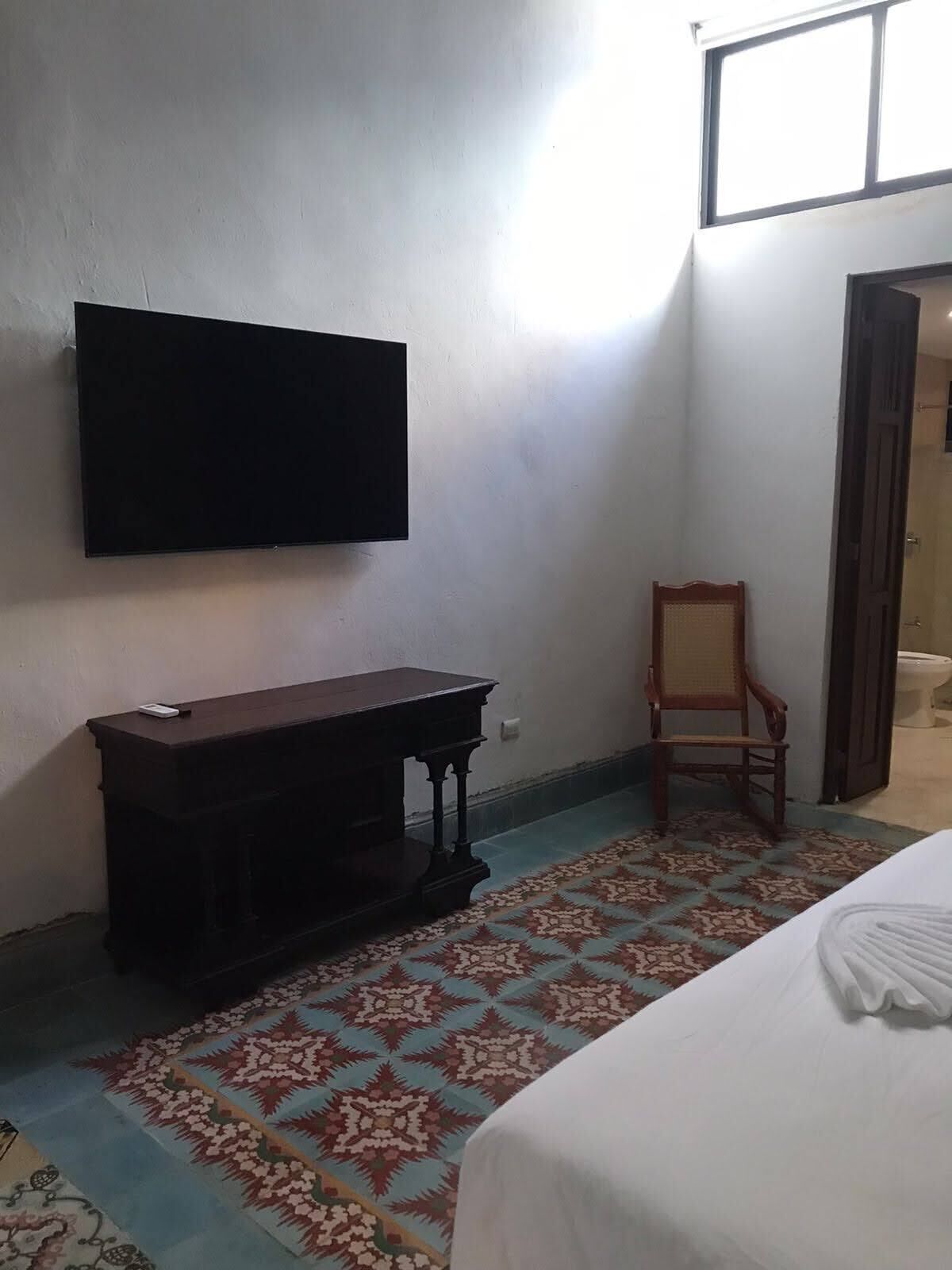 A room with a bed, a wooden bench, a chair, a television mounted on the wall, and a patterned rug on the floor.