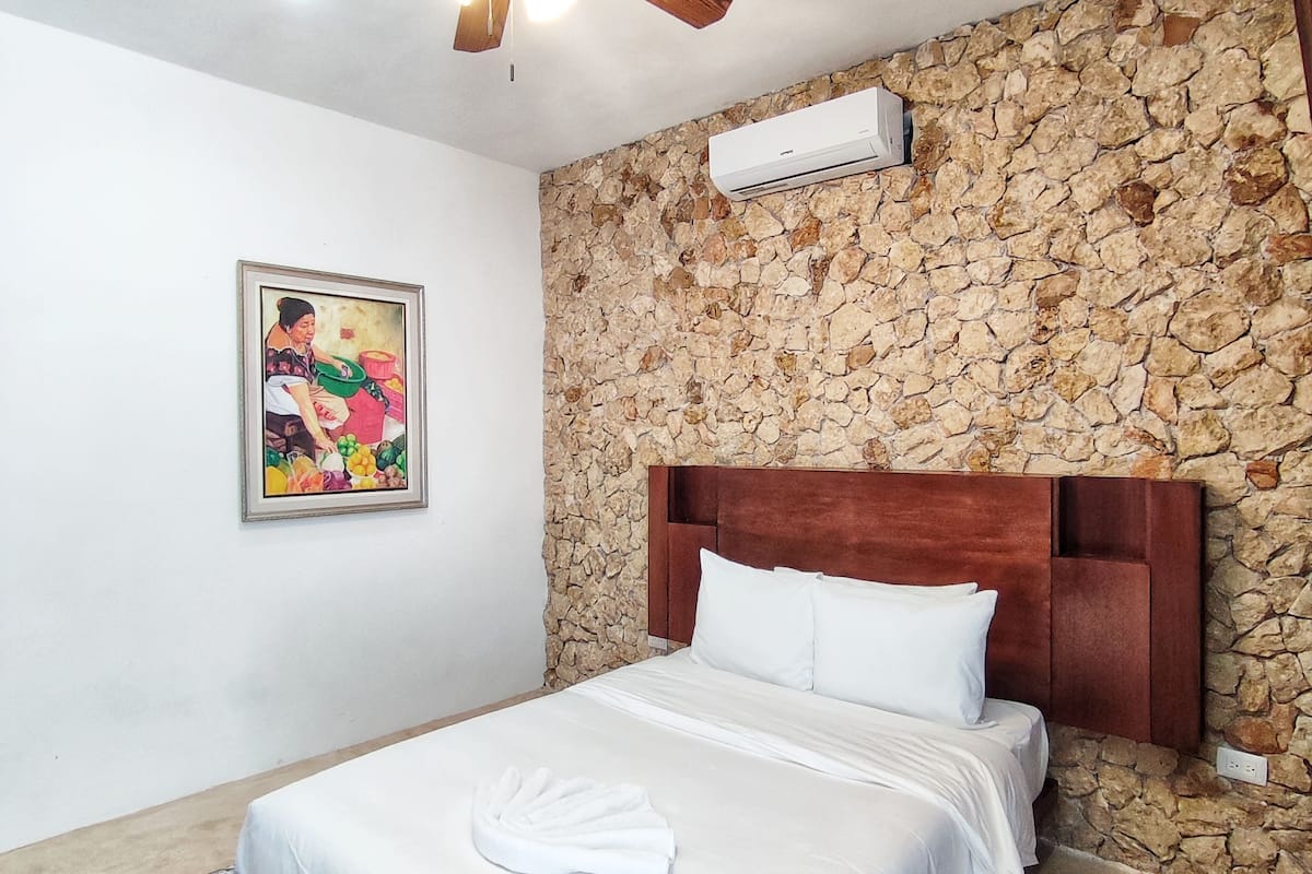 A bedroom with a bed, a ceiling fan, a stone wall, an air conditioning unit, and a painting.
