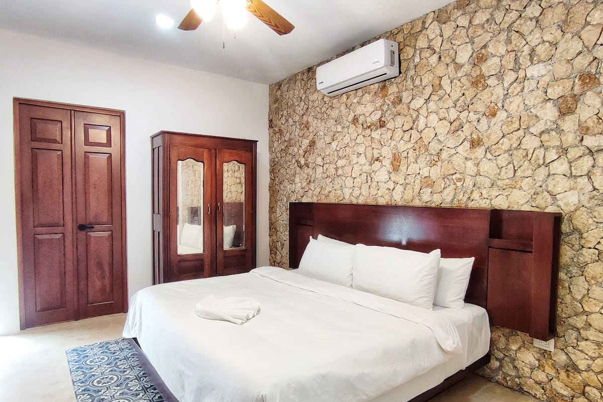 A bedroom with a bed, a ceiling fan, a stone wall, and an air conditioning unit.