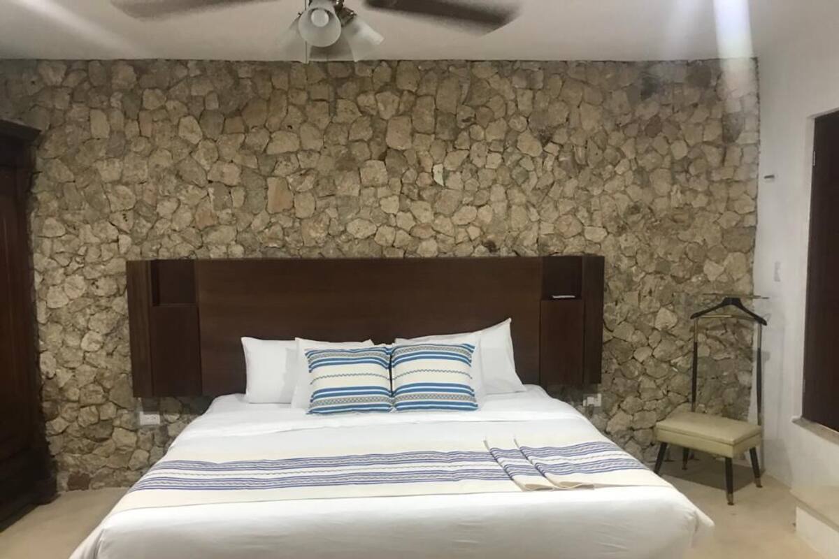 A bedroom with a stone wall, a wooden headboard, a bed with white bedding and striped pillows, and a small bedside table with a lamp.